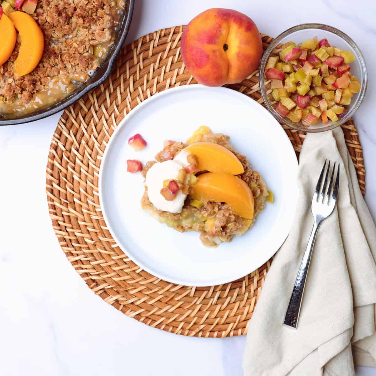 A plate of rhubarb crisp beside the skillet of dessert topped with iced cream and sliced peaches, displayed with a bowl of rhubarb, a whole peach a napkin and fork.