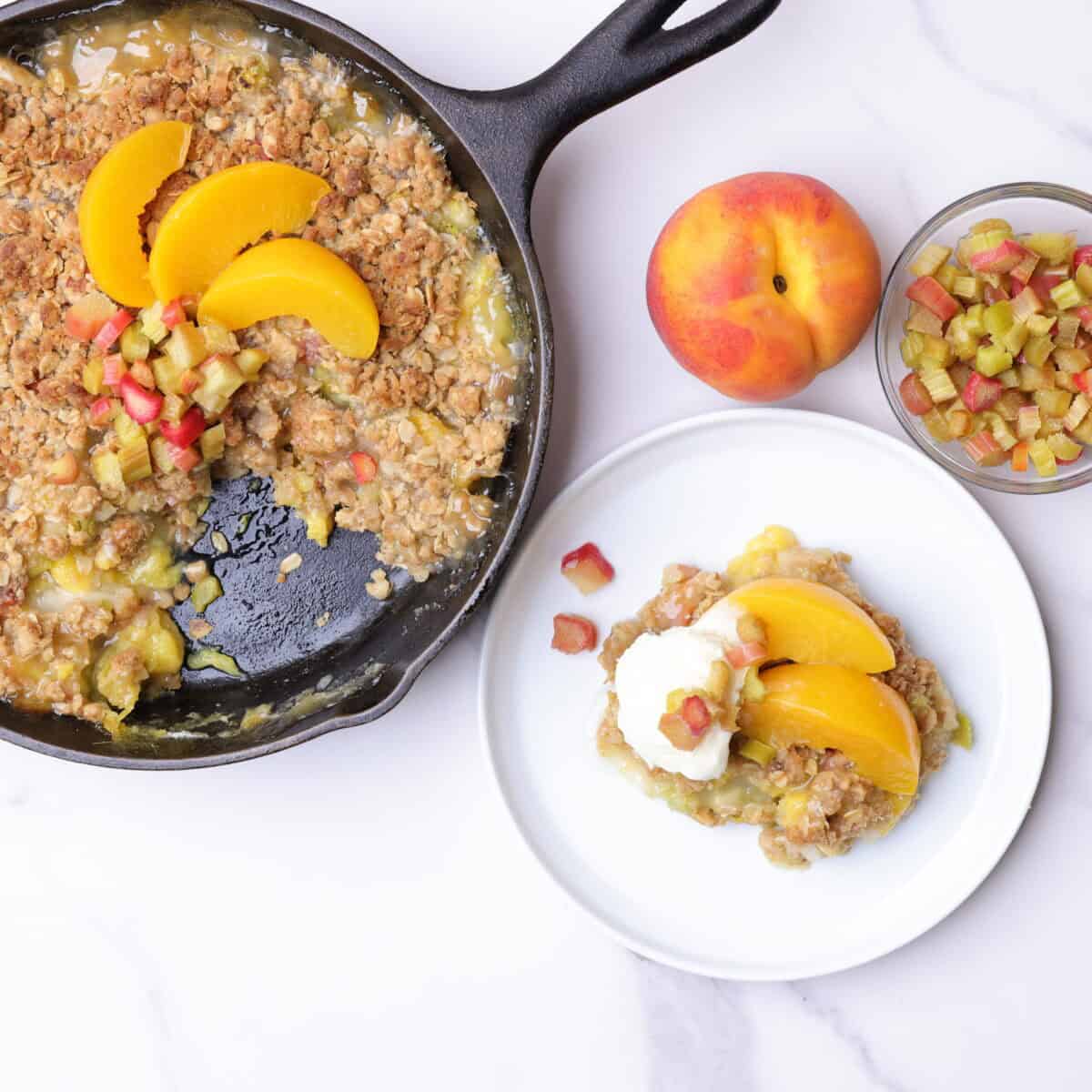 A skillet with rhubarb peach crisp topped with sliced rhubarb and peaches. A slice of crisp removed and plated on a white plate, topped with iced cream and peaches, displayed with a peach and bowl of sliced rhubarb.