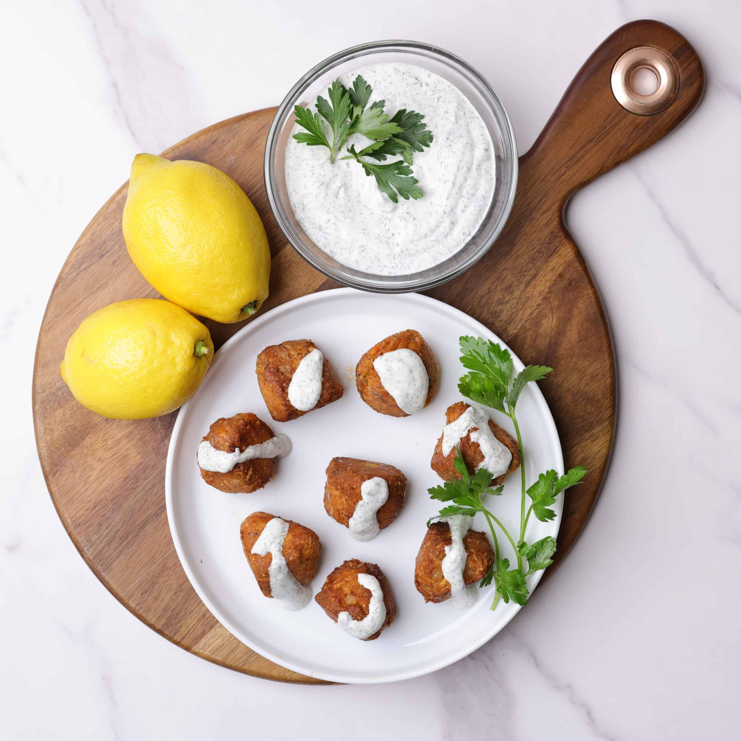 A round plate with salmon meatballs arranged on it, topped with dill sauce, garnished with parsley and displayed on a charcuterie board with lemons and a bowl of dill sauce. 