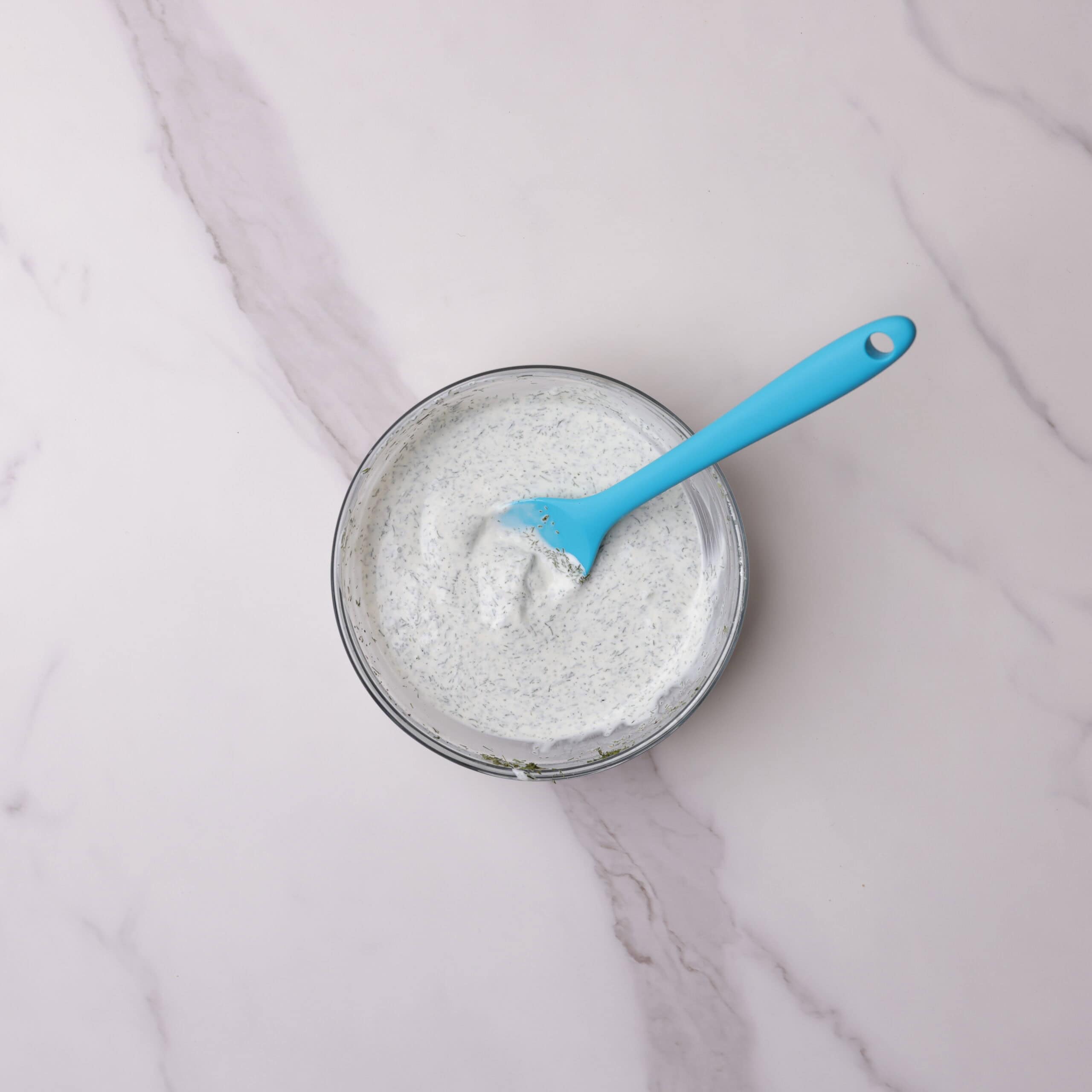 A small glass bowl the the dill sauce mixed together with a blue spatula. 