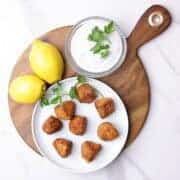 A round plate with salmon meatballs arranged on it, garnished with parsley and displayed on a charcuterie board with lemons and a bowl of dill sauce.