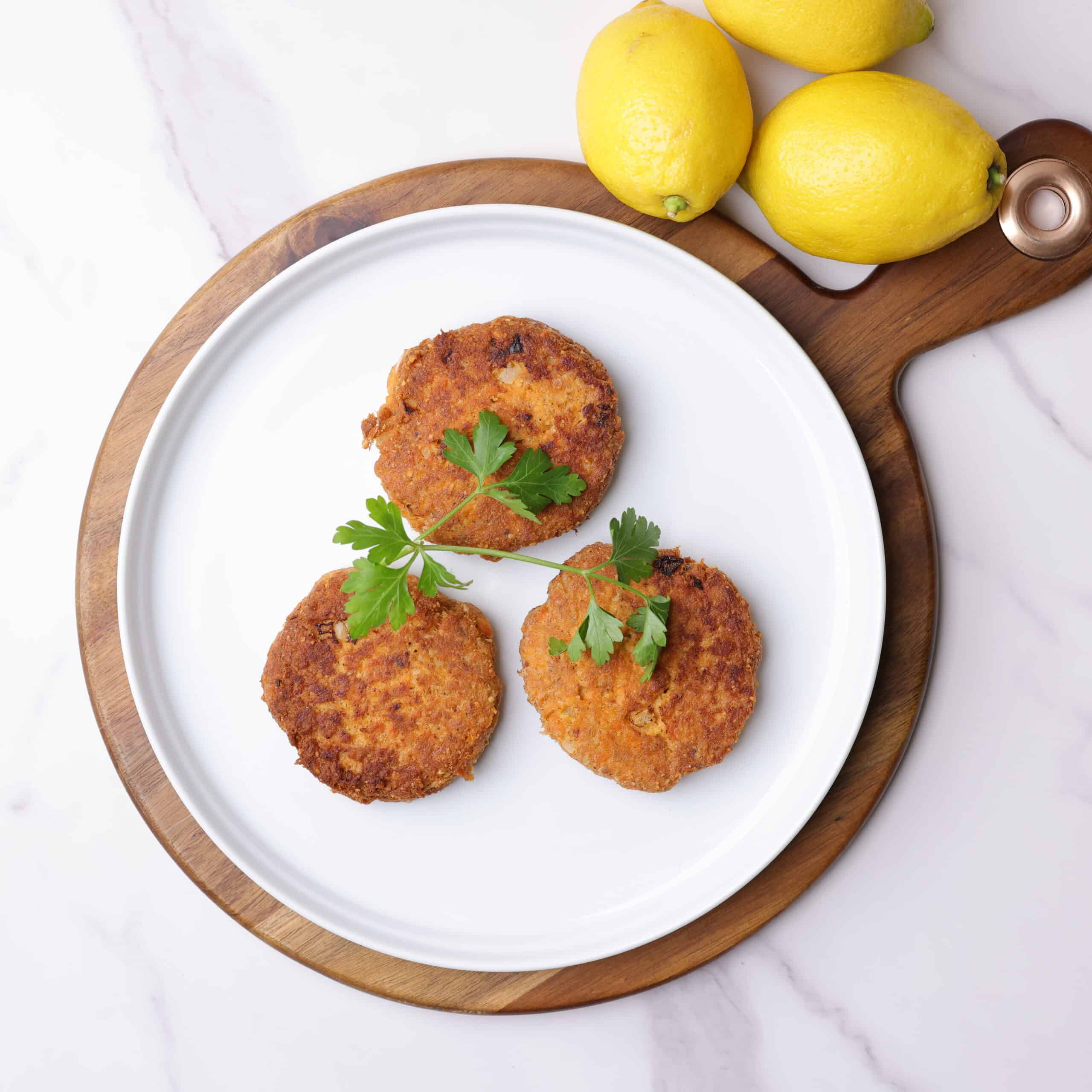 Three cooked salmon patties on a white plate garnished with parsley and displayed on a charcuterie board with lemons.