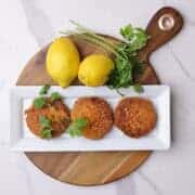 Three salmon patties plated on a white rectangular plate displayed on a charcuterie board with lemons and parsley beside.