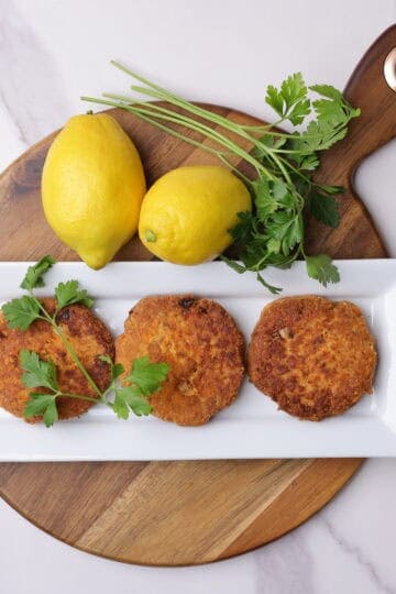Three salmon patties plated on a white rectangular plate displayed on a charcuterie board with lemons and parsley beside.