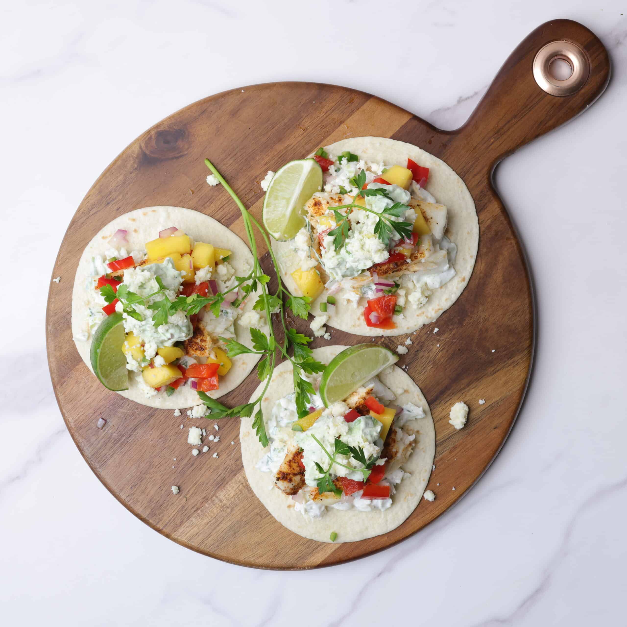 Three fully assembled, open faced blackened halibut tacos on a wooden charcuterie board, garnished with parsley and lime wedges. 