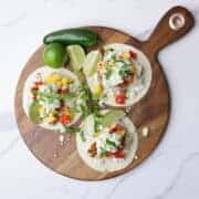 Three open faced, fully assembled blackened halibut tacos on a wooden charcuterie board, embellished with limes and a jalapeno pepper.