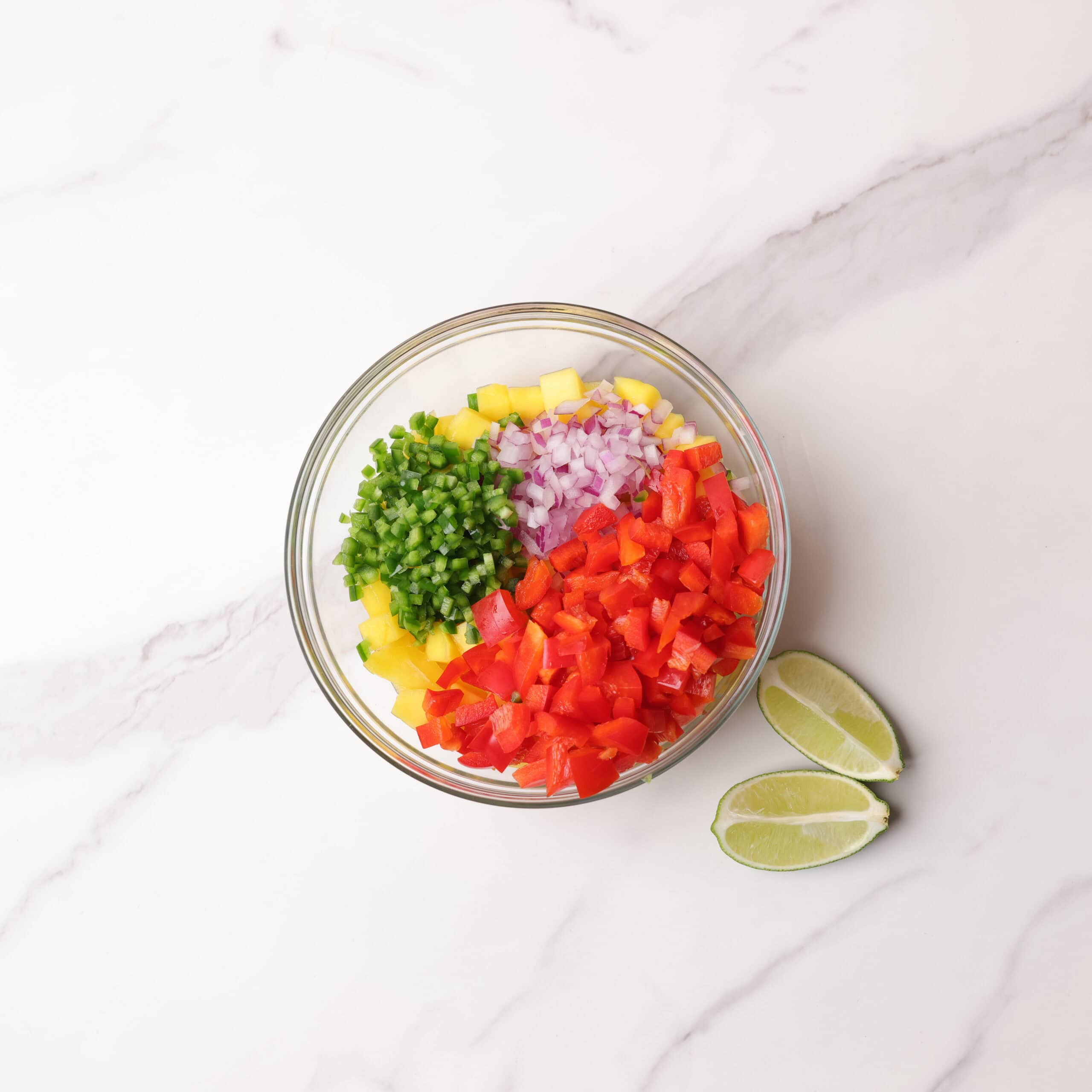 A glass bowl with chopped mango, purple onion, red bell pepper, jalapeno and lime wedges on the side. 