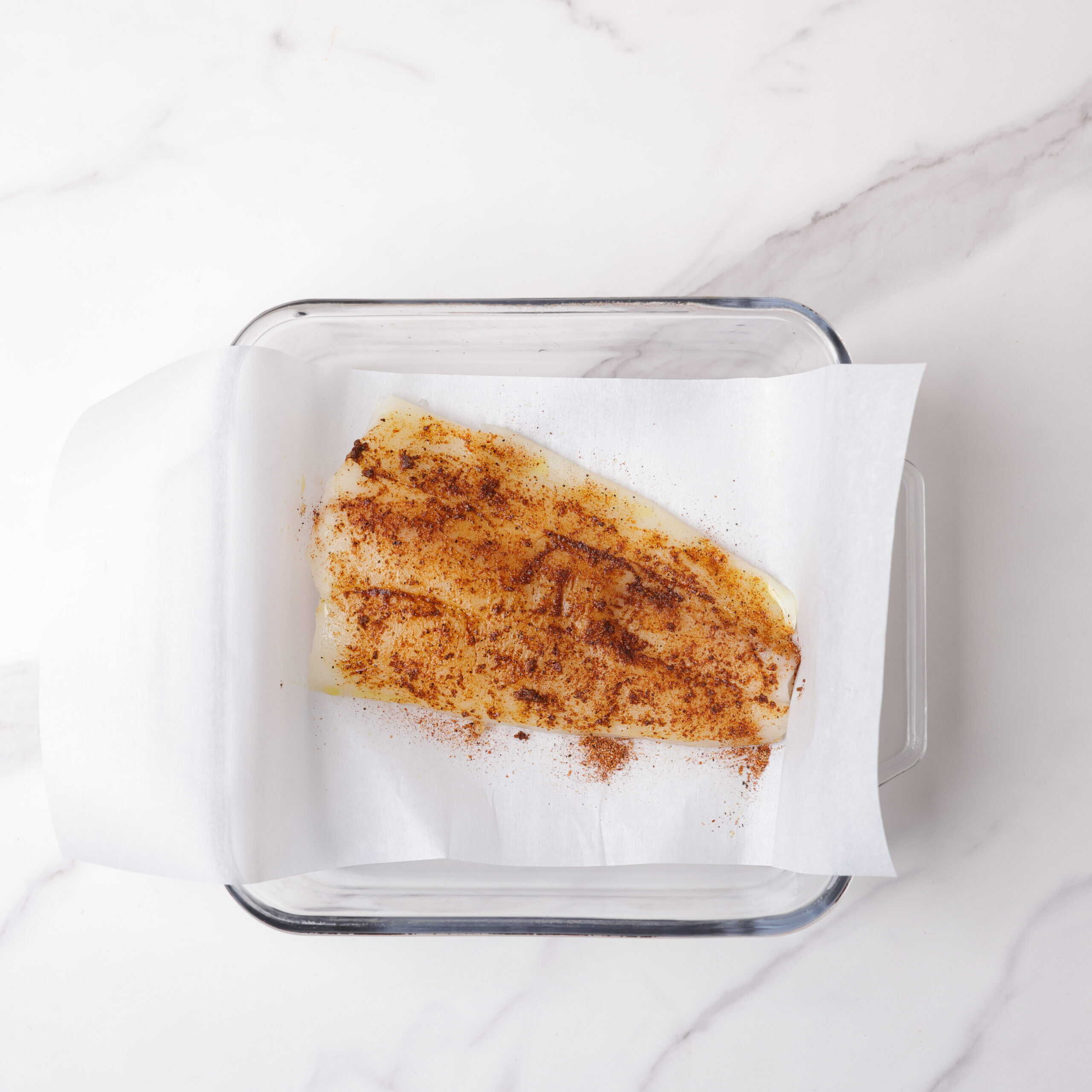 A halibut fillet oiled and seasoned in a glass baking dish on a piece of parchment paper. 