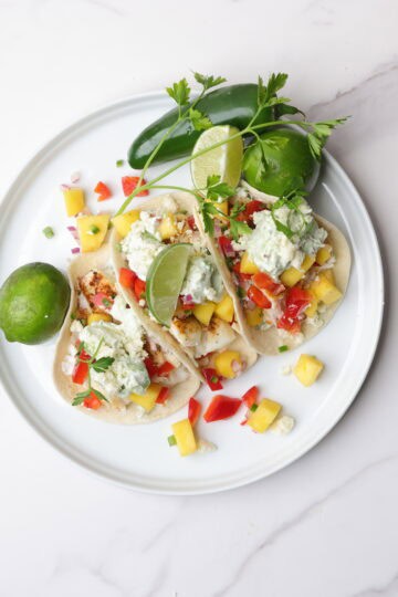 Three Blackened Halibut Tacos displayed on a white plate with limes, a jalapeno and parsley.