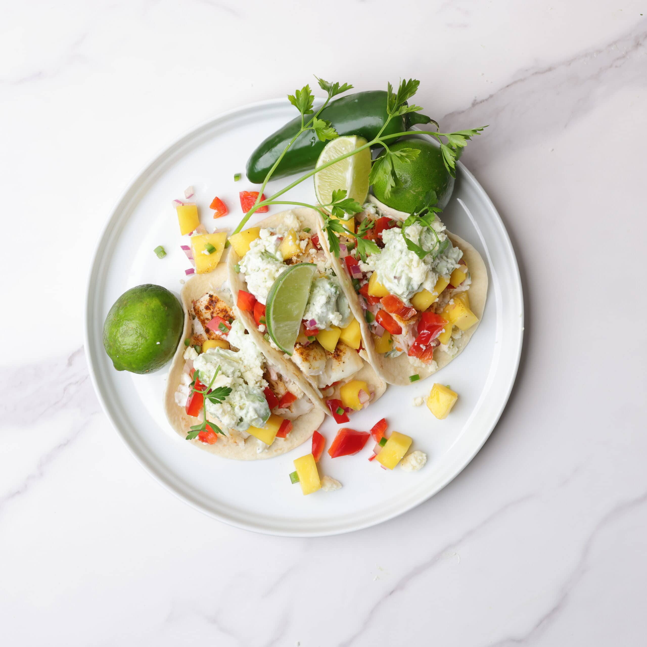 Three Blackened Halibut Tacos displayed on a white plate with limes, a jalapeno and parsley.