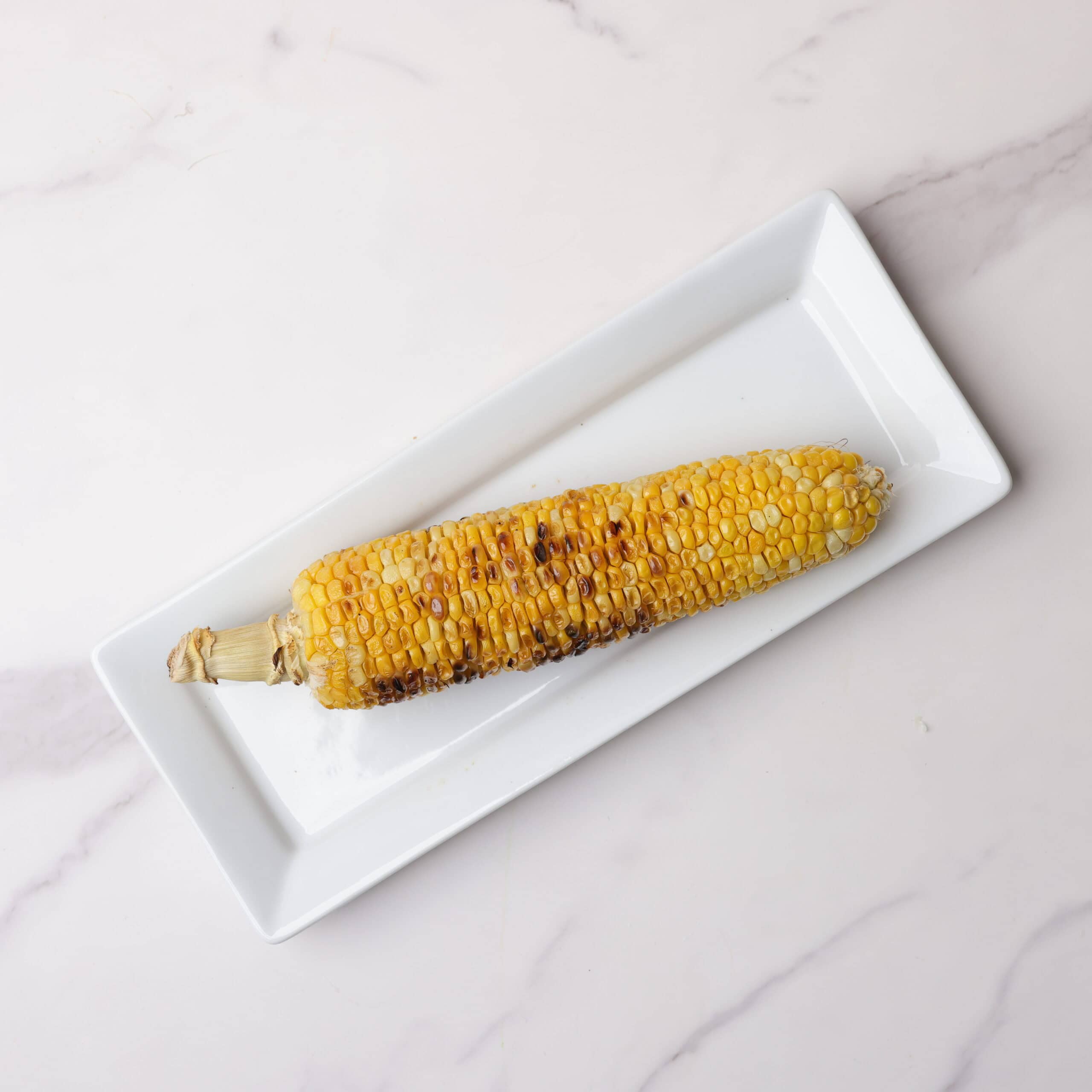 A charred cob of corn displayed on a white rectangular plate.