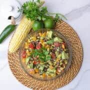 A large glass bowl filled with charred corn salad, displayed on a wicker place mat with a cob of corn, jalapeno, lime and jar of honey alongside and garnished with parsley.
