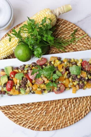 A white rectangular plate filled with charred corn salad, displayed on a wicker place mat with a cob of corn, jalapeno, lime and jar of honey alongside and garnished with parsley.