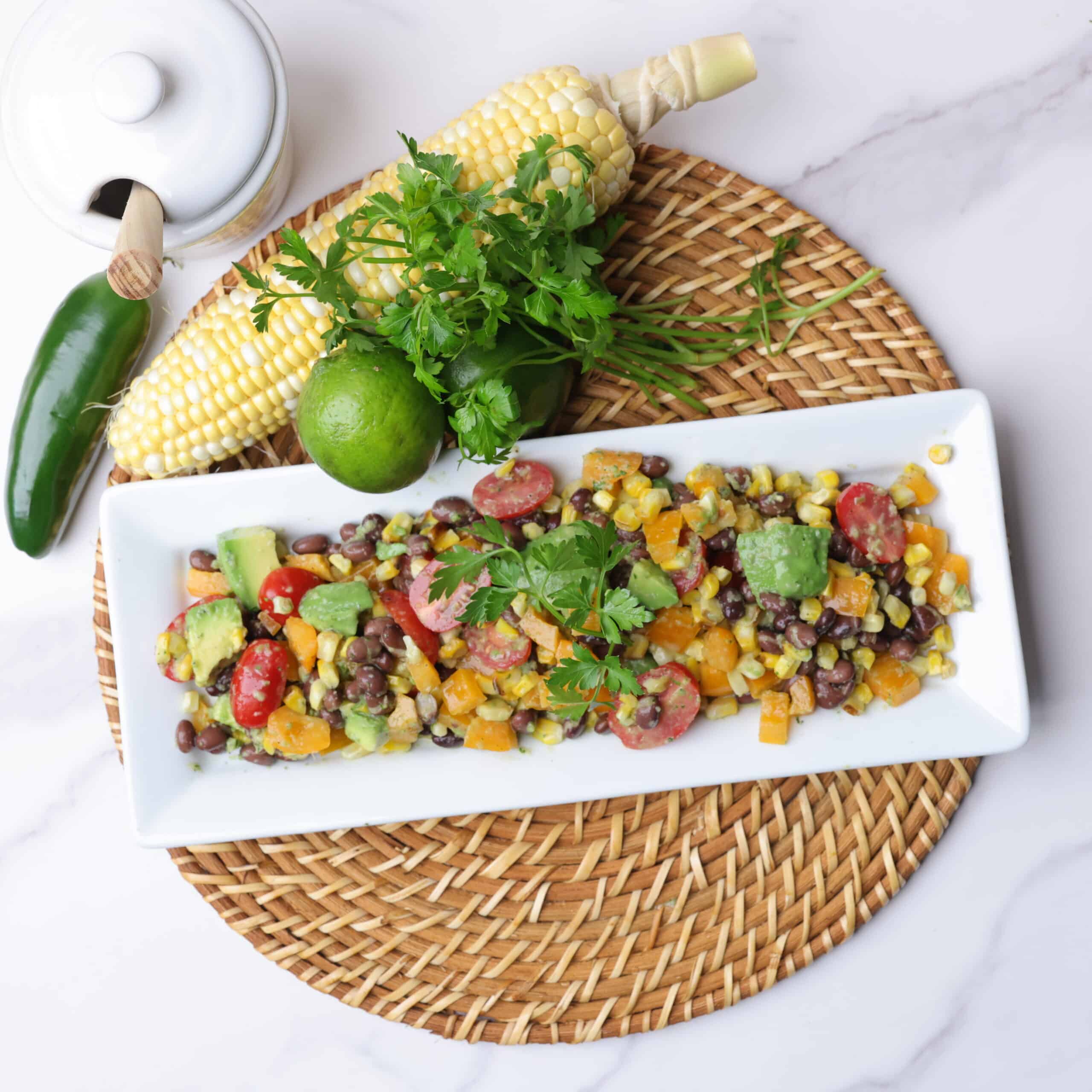 A white rectangular plate filled with charred corn salad, displayed on a wicker place mat with a cob of corn, jalapeno, lime and jar of honey alongside and garnished with parsley.