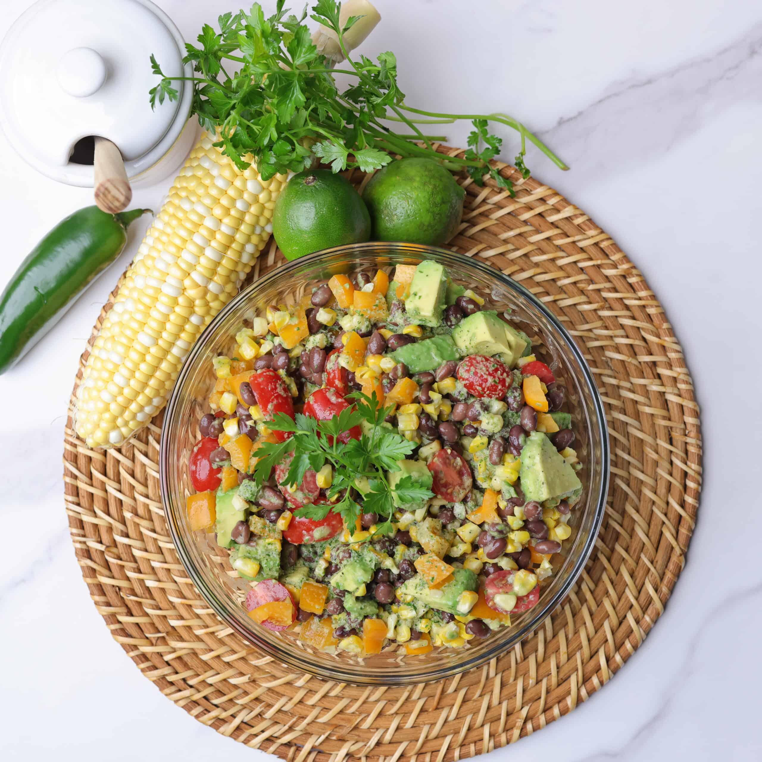 A large glass bowl filled with charred corn salad, displayed on a wicker place mat with a cob of corn, jalapeno, lime and jar of honey alongside and garnished with parsley.