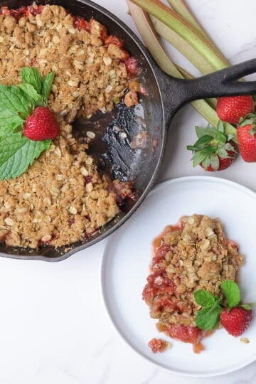 Strawberry Rhubarb Crisp baked in a cast iron skillet and displayed with a piece removed and plated on a white plate, both garnished with mint leaves and strawberries with sticks of rhubarb and strawberries on the side.