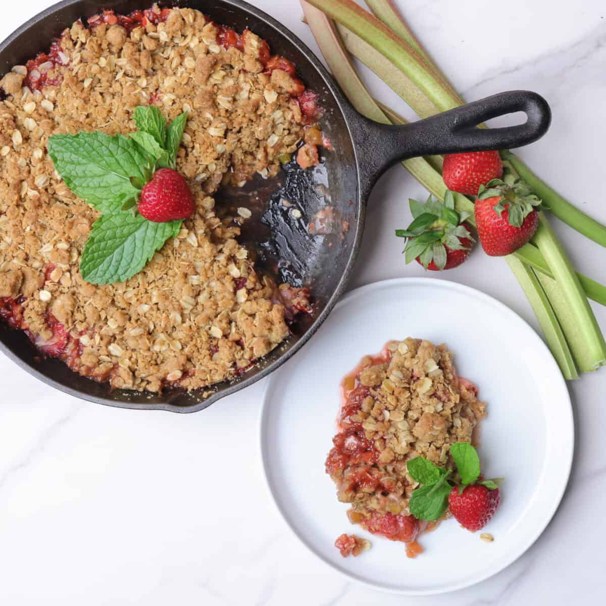 Strawberry Rhubarb Crisp baked in a cast iron skillet and displayed with a piece removed and plated on a white plate, both garnished with mint leaves and strawberries with sticks of rhubarb and strawberries on the side. 