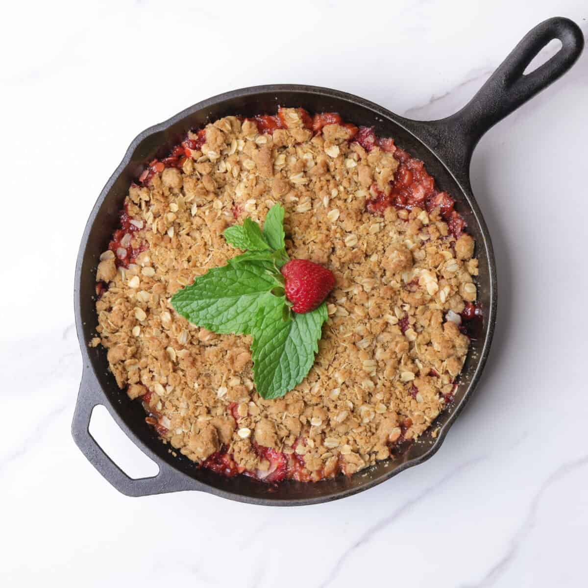 A cast iron skillet with a golden brown bake crisp garnished with mint leaves and a strawberry.