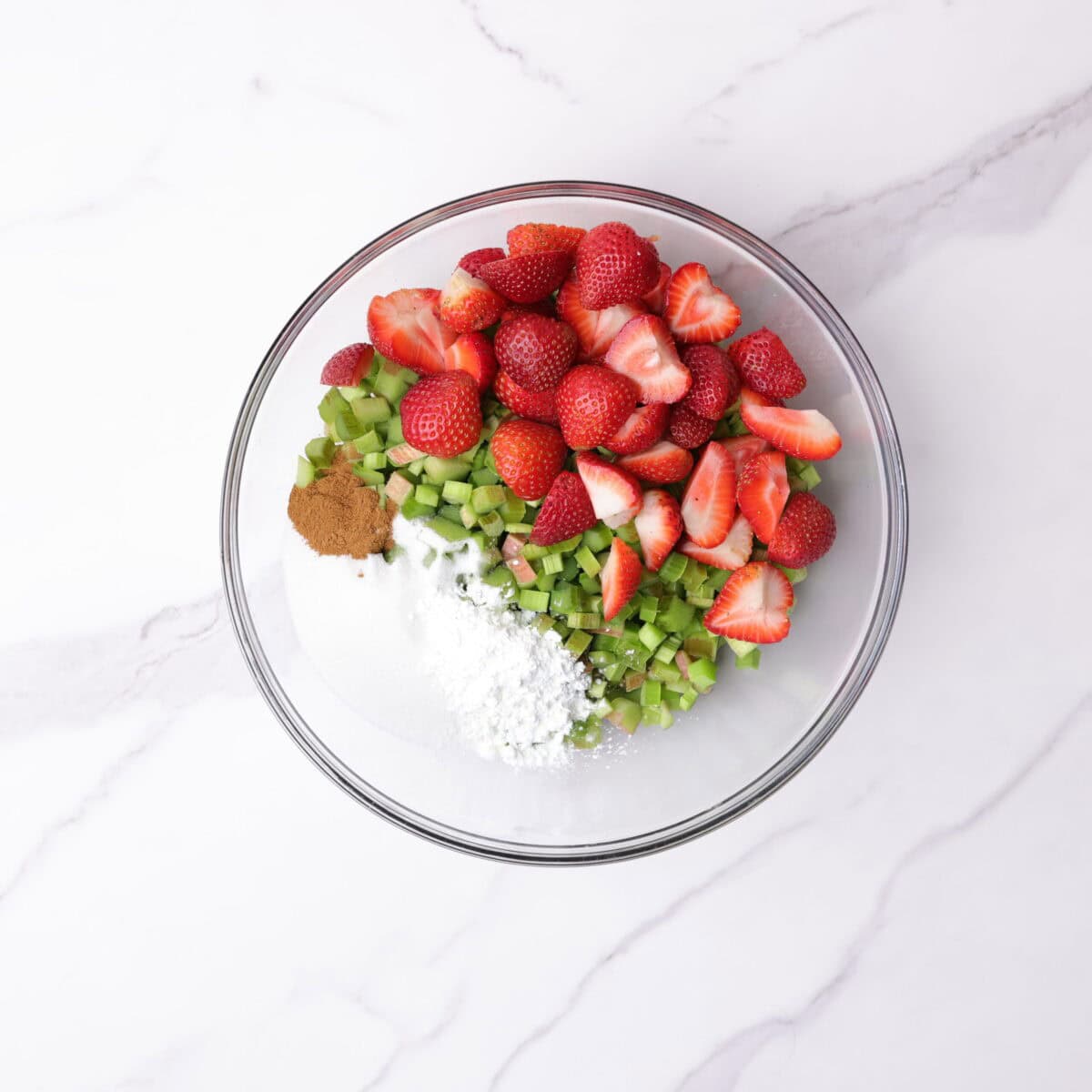 A large glass bowl containing the chopped rhubarb and strawberries, granulated sugar, cornstarch, cinnamon and vanilla.