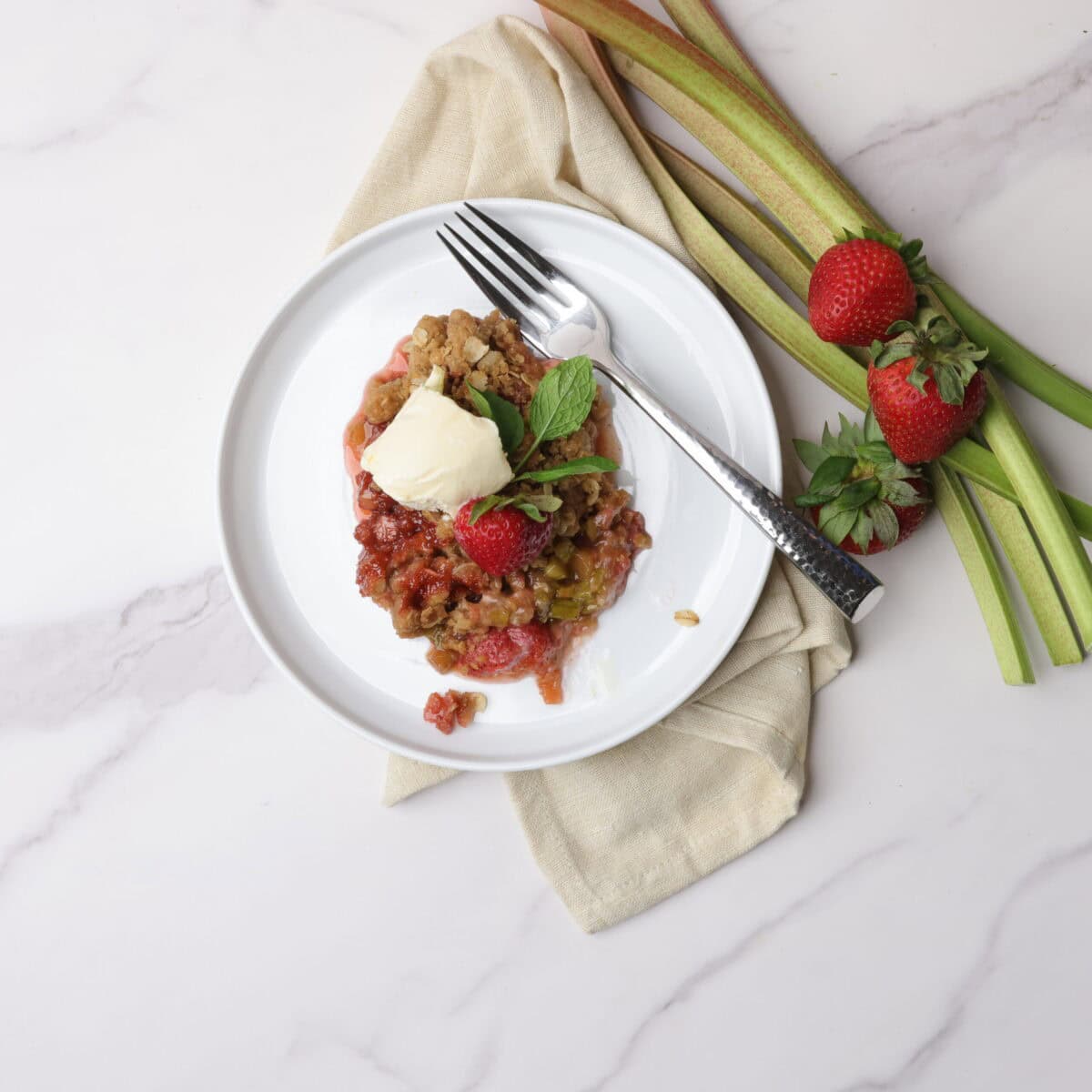 A white plate and fork holding a scoop of strawberry rhubarb crisp topped with ice cream, garnished with a strawberry and mint leaves, rests on a napkin with rhubarb sticks and strawberries beside it.