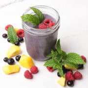 A mason jar filled with blueberry smoothie, garnished with mint leaves and raspberries and surrounded by mangoes, blueberries, raspberries and mint leaves for display.