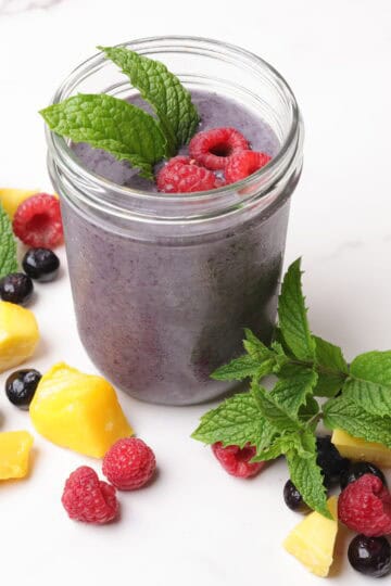 A mason jar filled with blueberry smoothie, garnished with mint leaves and raspberries and surrounded by mangoes, blueberries, raspberries and mint leaves for display.