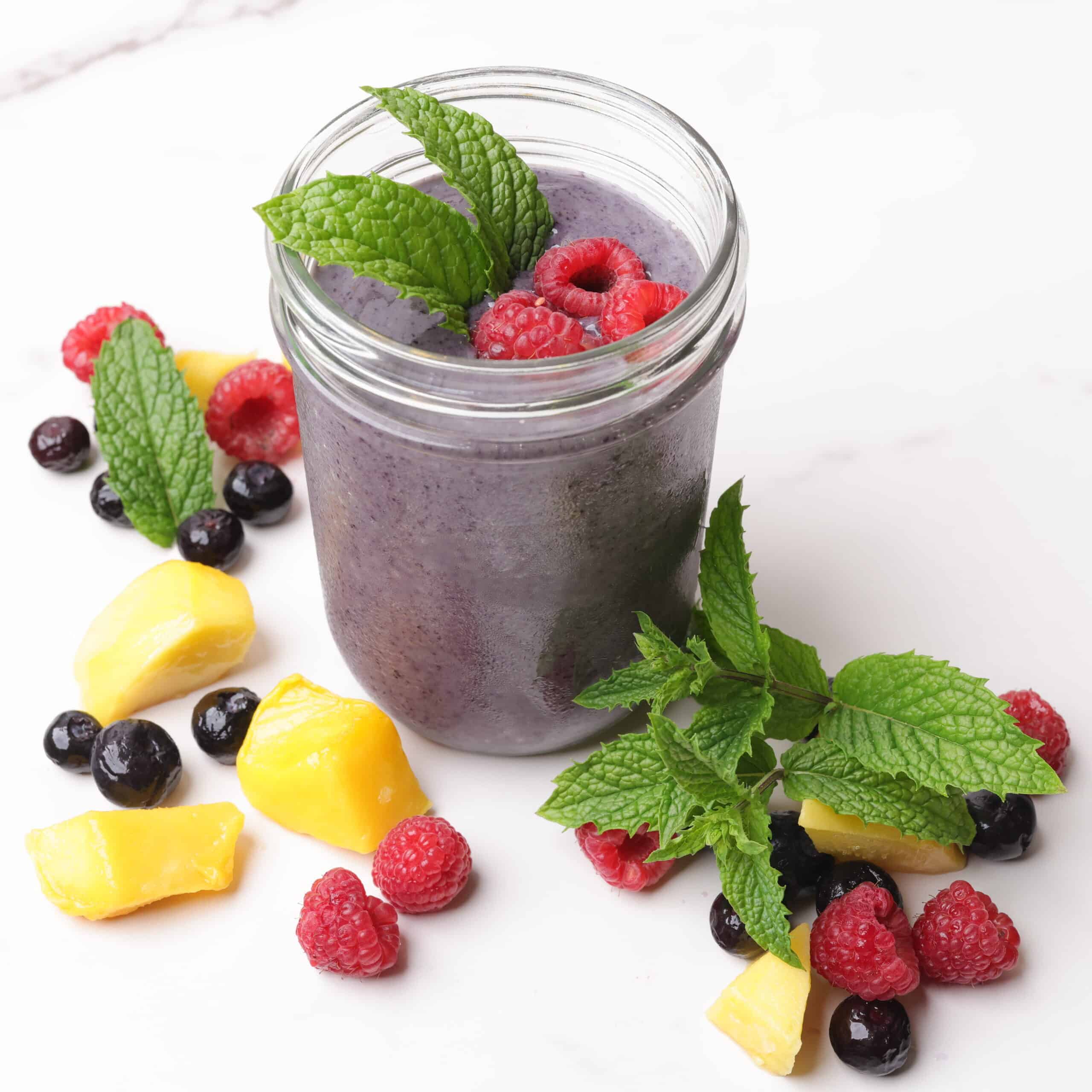 A mason jar filled with blueberry smoothie, garnished with mint leaves and raspberries and surrounded by mangoes, blueberries, raspberries and mint leaves for display.