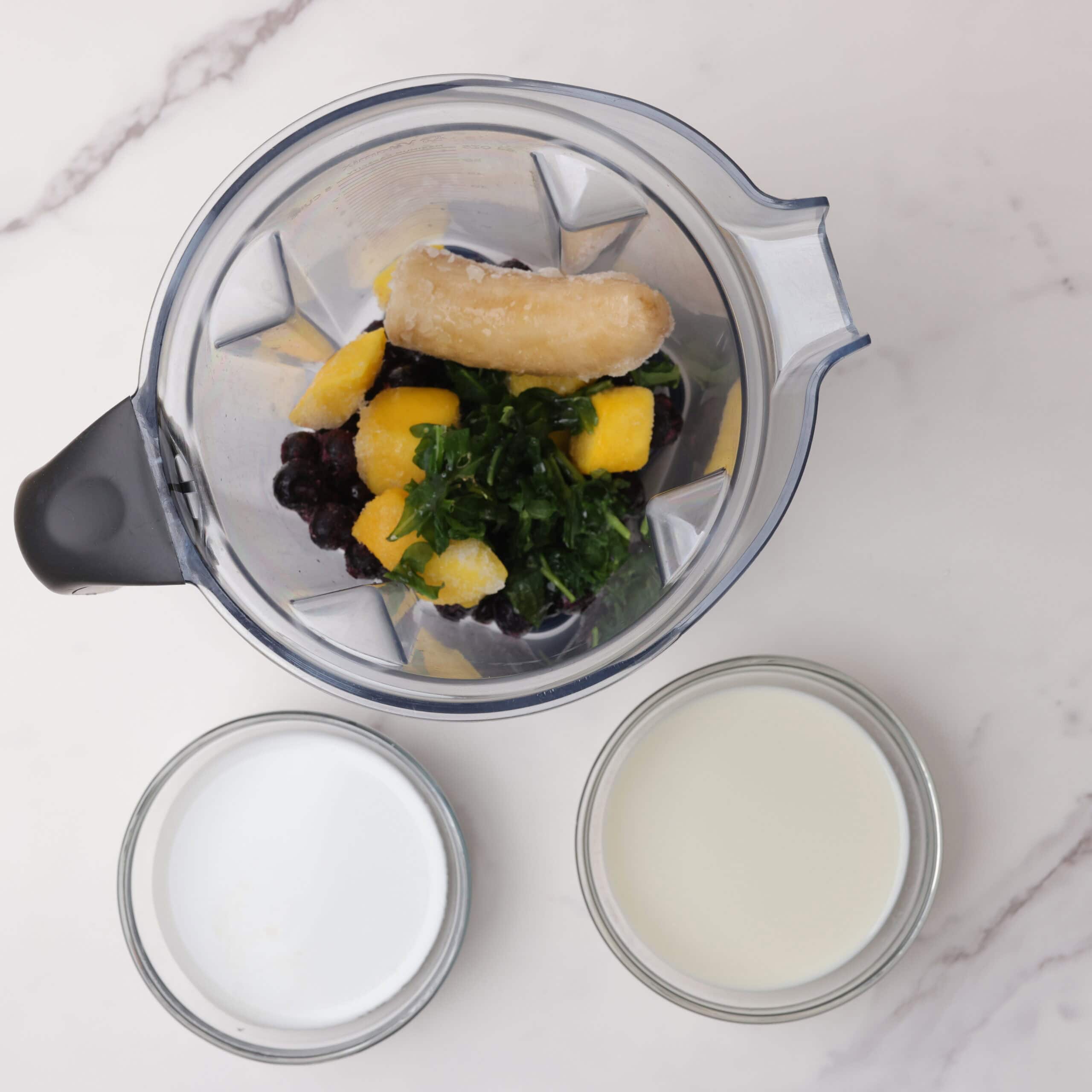 A vitamix with the frozen blueberries, mangoes, banana and kale in it with bowls of the almond and coconut milk beside it.
