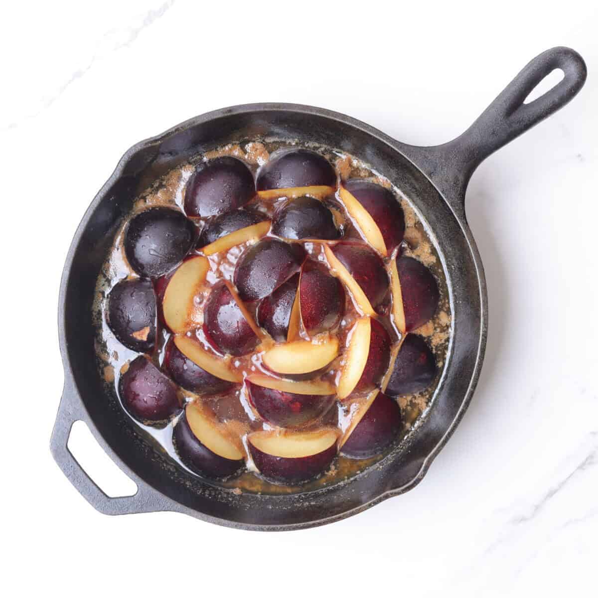 The skillet with the butter and sugar mixture melted, now with quartered plums arranged in circle form in the pan, creating a flower like pattern.