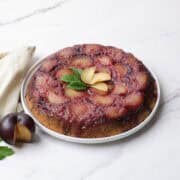 The upside down plum cake inverted on the white serving plate, garnished with fresh plums and mint leaves and displayed with a sliced plum and cloth napkin beside.