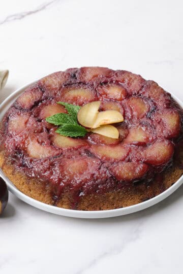 The upside down plum cake inverted on the white serving plate, garnished with fresh plums and mint leaves and displayed with a sliced plum and cloth napkin beside.
