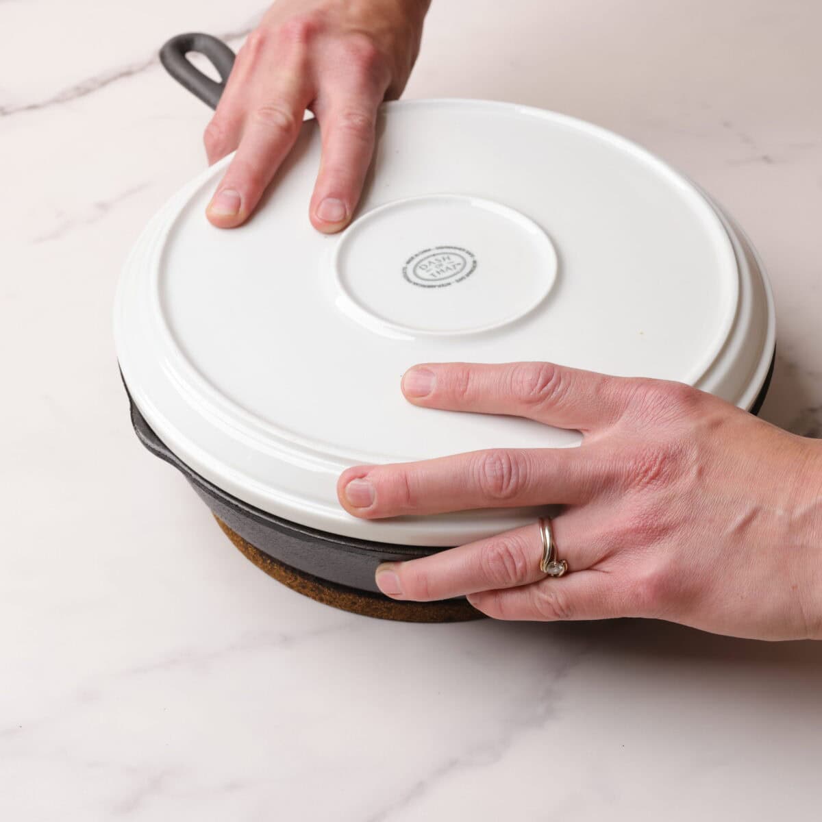 The skillet with the baked plum upside down cake in it with a white plate over it with two hands holding the plate in place.