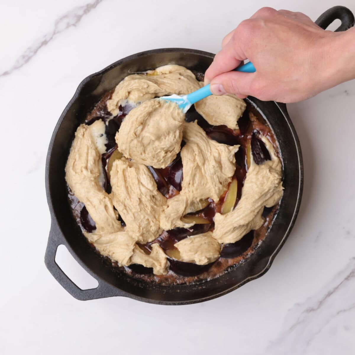 A hand spooning the batter over the cooked plums in the skillet.