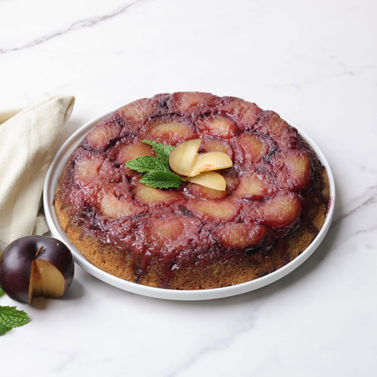 The plum upside down cake inverted on the white serving plate, garnished with fresh plums and mint leaves and displayed with a sliced plum and cloth napkin beside.