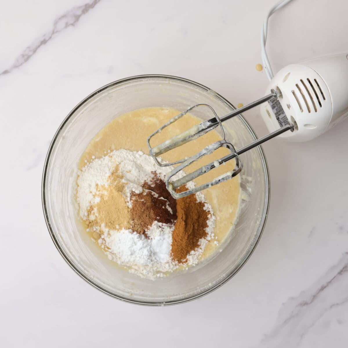 A glass bowl with the batter mixture topped with the dry ingredients and a hand mixer resting on the edge.