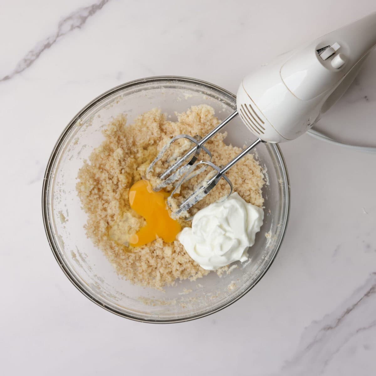 A glass bowl with the blended butter and sugar topped with the egg and yogurt with a hand mixer resting on the edge.