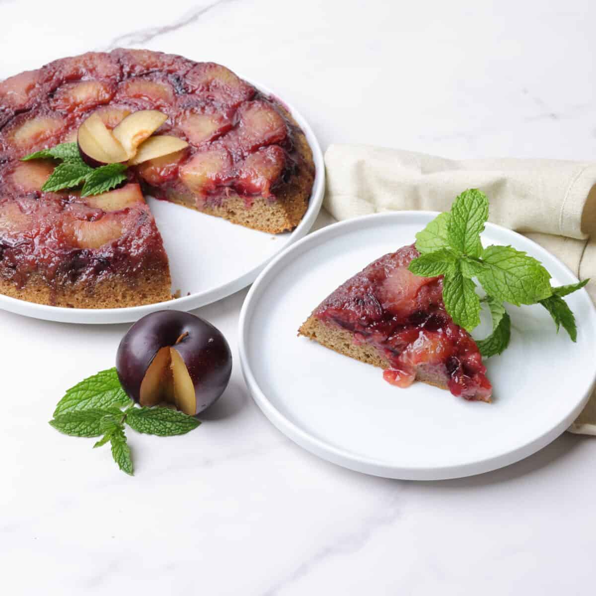 Plum upside down cake on a serving plate with a slice taken out and displayed on a smaller white plate. All displayed with a sliced plum, mint leaves and a napkin beside.