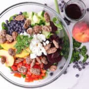 A glass bowl of Peach and Blueberry Salad garnished with parsley and with a peach, blueberries, bundle of parsley and cup of blueberry balsamic vinaigrette beside it.