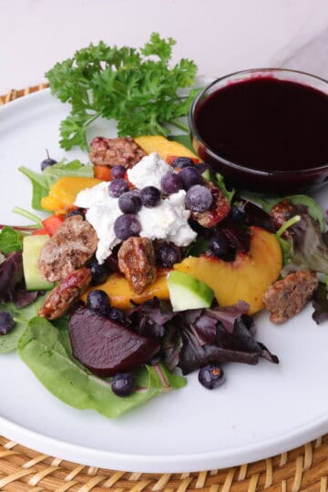 Peach and Blueberry Salad displayed on a white plate with a bowl of blueberry balsamic vinaigrette and parsley for garnish.