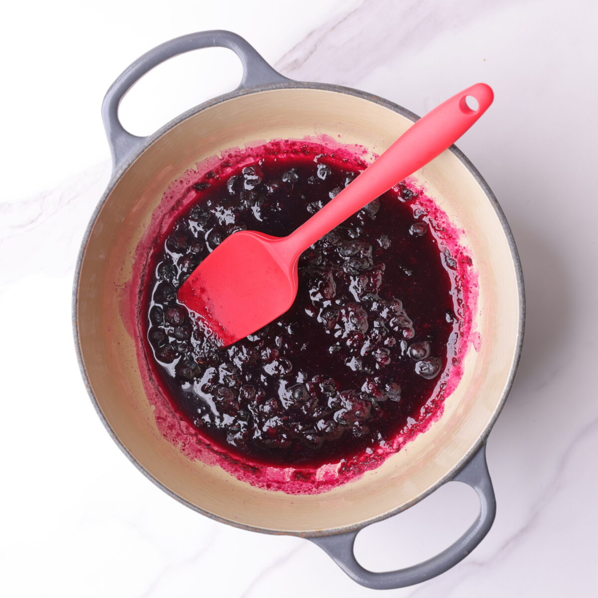 A small pot with blueberries cooked down and a red spatula in it for stirring.