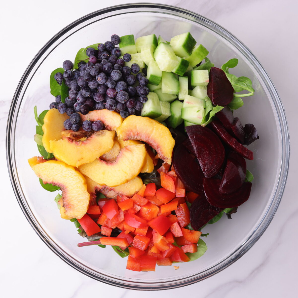 A glass bowl with mixed greens and the blueberries, peaches, cucumbers, bell peppers and beets arranged on top.