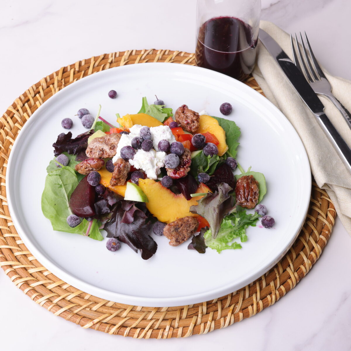 A white plate with a portion of the Peach and Blueberry Salad arranged on it, sitting on a wicker place mat with cutlery and a napkin beside it and a jar of blueberry balsamic vinaigrette above. 