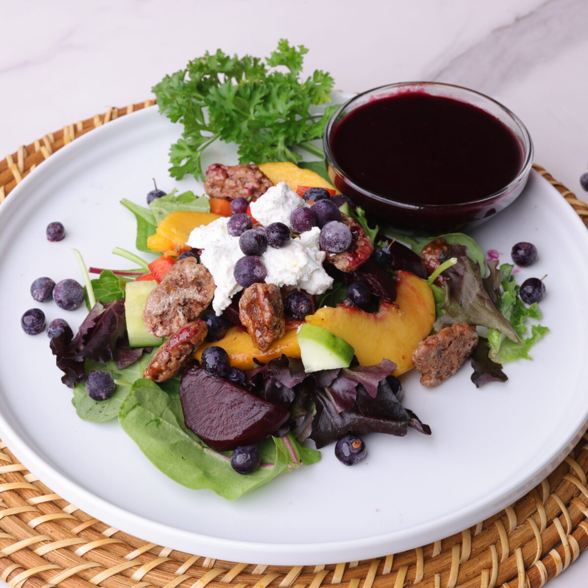 Peach and Blueberry Salad displayed on a white plate with a bowl of blueberry balsamic vinaigrette and parsley for garnish.