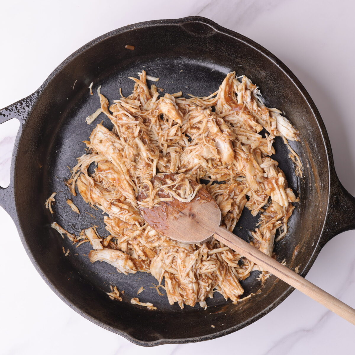 A raw cast iron skillet with shredded chicken coated in barbecue sauce with a wooden spoon in it for stirring.