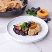 Blueberry Peach Cobbler displayed on a white plate and garnished with mint leaves and fresh blueberries with mint, blueberries and a half peach beside it and the remaining cobbler in the skillet in the background.