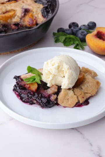 Blueberry Peach Cobbler displayed on a white plate and garnished with mint leaves and ice cream with mint, blueberries and a half peach beside it and the remaining cobbler in the skillet in the background.