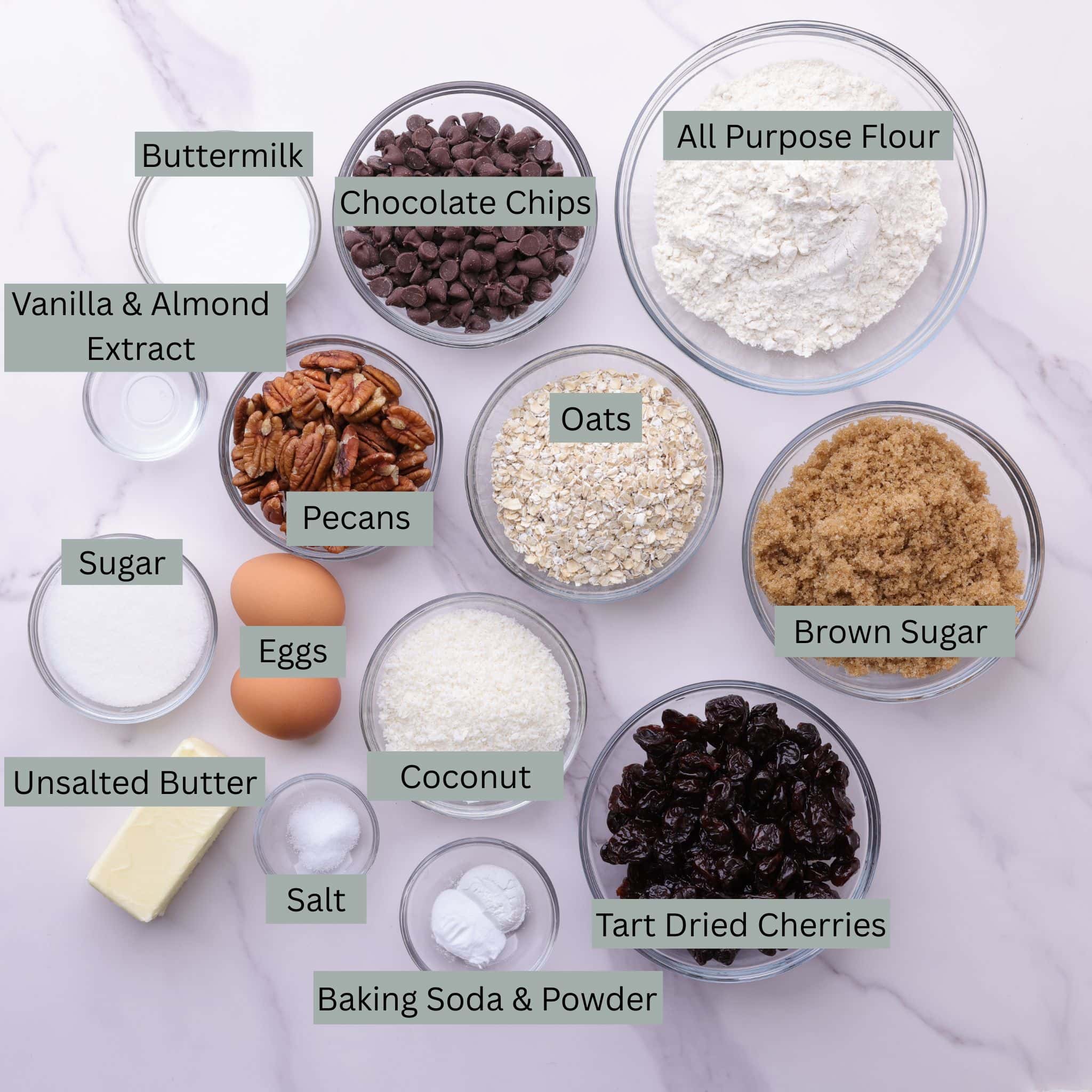 A visual display of the ingredients for the Oatmeal Cherry Cookies - buttermilk, chocolate chips, flour, vanilla and almond extract, pecans, oats, brown sugar, granulated sugar, eggs, coconut, dried tart cherries, butter, salt, baking soda and baking powder - each in their own bowl.