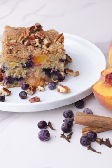 A slice of Peach and Blueberry Coffee Cake displayed on a white plate with peaches, blueberries and cinnamon sticks beside.