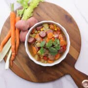 A bowl of Sausage and Pepper Soup displayed on a wooden board with carrots, celery and potatoes beside it.