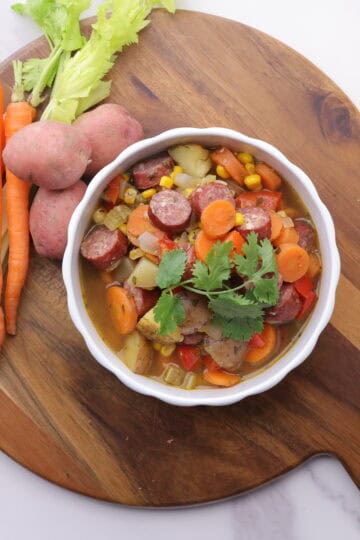 A bowl of Sausage and Pepper Soup displayed on a wooden board with carrots, celery and potatoes beside it.